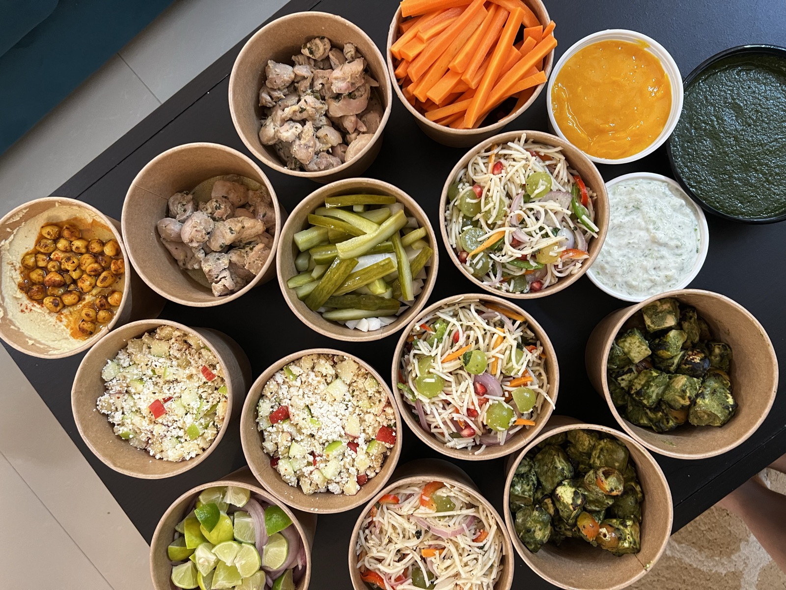 Overhead view of many kraft bowls filled with Pooja's catering prep — salads, tikka, chutneys and more
